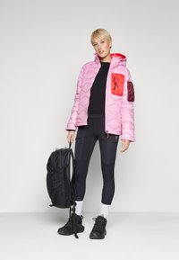 Gevoerde roze jas met rode en kastanjebruine accenten, zwarte legging, zwarte schoenen en een zwarte rugzak. Heeft een gewatteerd ontwerp en capuchon.