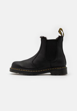 Bottes Chelsea en cuir noir avec surpiqûres jaunes, panneau latéral élastique, doublure en fourrure douce en haut, et languette de traction brandée à l'arrière.