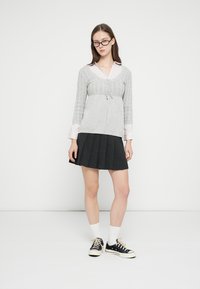 Pilkas megztinis su siūlėta tekstūra ir raišteliu juosmenyje, dėvimas ant šviesiai rožinės palaidinės su apykakle; dera su juoda klostuota suknele ir sportbačiais.