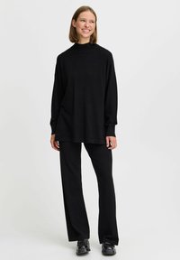 Bluză neagră supradimensionată cu mâneci lungi, guler înalt și pantalonii negri asortați cu croială largă. Textură fină; design minimal, fără modele vizibile.