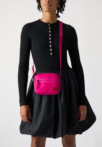 Borsa a tracolla rosa con una texture liscia, compartimenti con zip e una tracolla lavorata, indossata su un top nero a costine e una gonna.