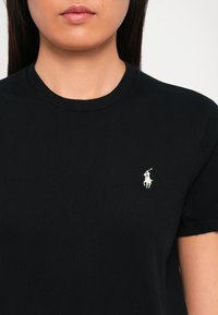Frau trägt ein eng anliegendes schwarzes Kurzarmshirt mit einem kleinen weißen Polo-Spieler-Logo, das auf der linken Brust gestickt ist.