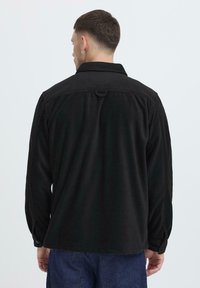 Black, long-sleeved shirt with a collar, buttoned cuffs, and a straight cut. Made of a soft, textured fabric, featuring a rear loop detail.