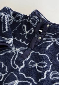 Pulover din fleece bleu marin cu un model de fundițe albe, având un guler înalt și butoane cu cap de tip snap. Material moale, texturat, cu margini contrastante.