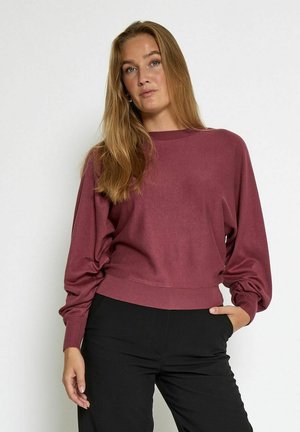 Maroon cropped sweater with long sleeves, ribbed cuffs, and a smooth texture, paired with black trousers. Simple design without embellishments.