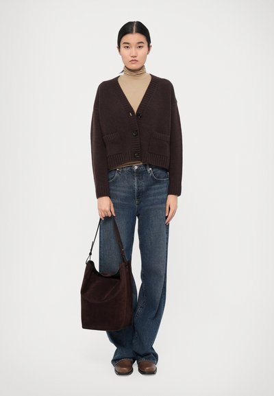 Brown knitted cardigan with large buttons, paired with a beige turtleneck and blue wide-leg jeans. A dark brown suede bag is held on one side.