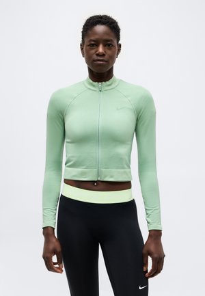 Femme portant une veste de sport zippée vert clair et des leggings noirs avec une ceinture vert clair, debout devant un fond uni.