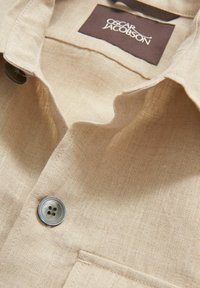 Beige linned skjorte med en foldet krave, en enkelt brystlomme og en fire-hulsknap med en iriserende finish. Mærket siger "Oscar Jacobson."
