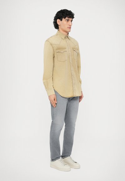 Chemise en denim beige clair avec deux poches poitrine et boutons pression, portée avec un jean gris clair et des baskets blanches. Coupe standard avec manches longues.