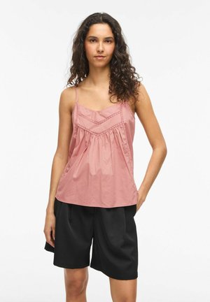 Femme aux longs cheveux bouclés portant un haut rose sans manches avec des détails en dentelle et un short noir ample, debout avec une main dans la poche.