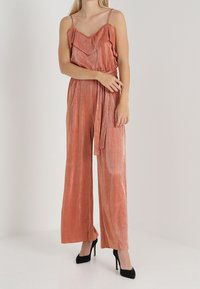 Mono en color rosa metálico con escote volado, cintura ceñida con cinturón y diseño de pierna ancha. Tirantes finos tipo espagueti y textura suave.