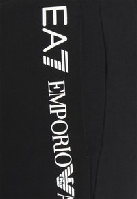 Černé teplákové kalhoty s bílým brandingem "EA" a "EMPORIO ARMANI" na boku. Vyrobené z hladkého, jemného materiálu s uvolněným střihem.