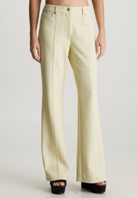 Pantalones acampanados de color amarillo claro con bolsillos frontales, detalles de costura centralizada y herrajes plateados, combinados con sandalias de plataforma negras.