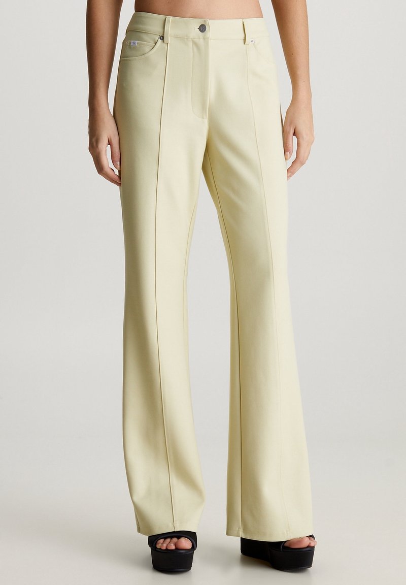 Pantalones acampanados de color amarillo claro con bolsillos frontales, detalles de costura centralizada y herrajes plateados, combinados con sandalias de plataforma negras.
