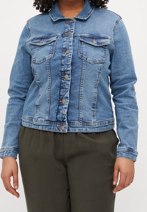 Frau trägt eine geknöpfte blaue Jeansjacke mit Brusttaschen und olivgrüner Hose vor einem schlichten Hintergrund.