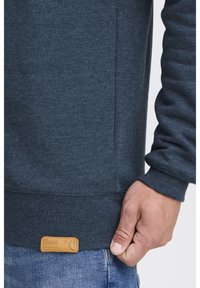 Gros plan d'une main reposant près de l'ourlet d'un sweat-shirt gris foncé avec une petite étiquette marron, et un jean en denim bleu visible.