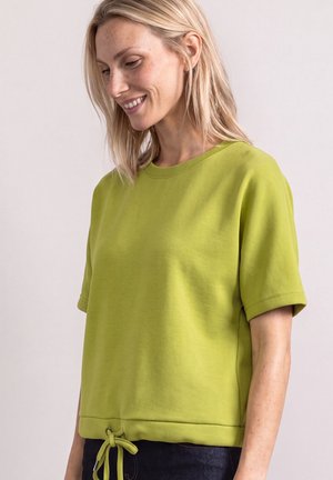 Femme aux cheveux blonds portant un haut vert citron à manches courtes avec un cordon à l’ourlet, souriante et regardant légèrement vers le bas sur le côté.