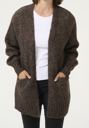 Personne portant un cardigan marron en maille avec des poches sur une chemise blanche et un pantalon noir, les mains dans les poches du cardigan.