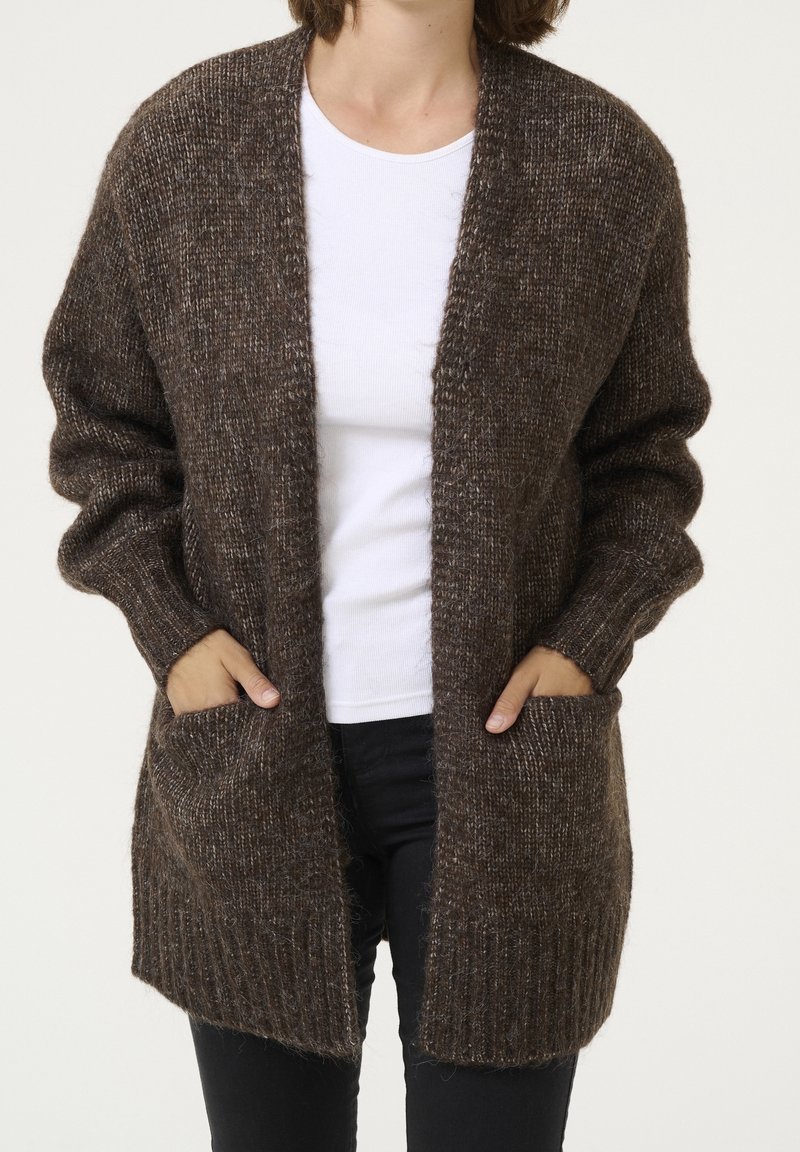 Personne portant un cardigan marron en maille avec des poches sur une chemise blanche et un pantalon noir, les mains dans les poches du cardigan.