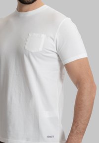 T-shirt bianco in cotone con colletto a giro, maniche corte e una tasca sul petto. Presenta un logo blu sul fondo dell'orlo.