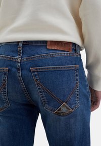 Jeans in denim blu scuro con una finitura testurizzata, caratterizzati da cuciture arancioni a contrasto, due tasche posteriori e un'etichetta in pelle sulla cintura.