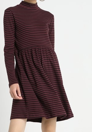 Frau trägt ein langärmliges, hochgeschlossenes burgunderrotes Kleid mit schwarzen horizontalen Streifen und einem gerafften Taillenbereich, steht vor einem weißen Hintergrund.