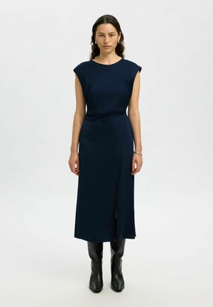 Donna in un vestito midi senza maniche di colore navy con uno spacco frontale, stivali neri fino al ginocchio e orecchini a cerchio in argento, in piedi davanti a uno sfondo bianco uniforme.