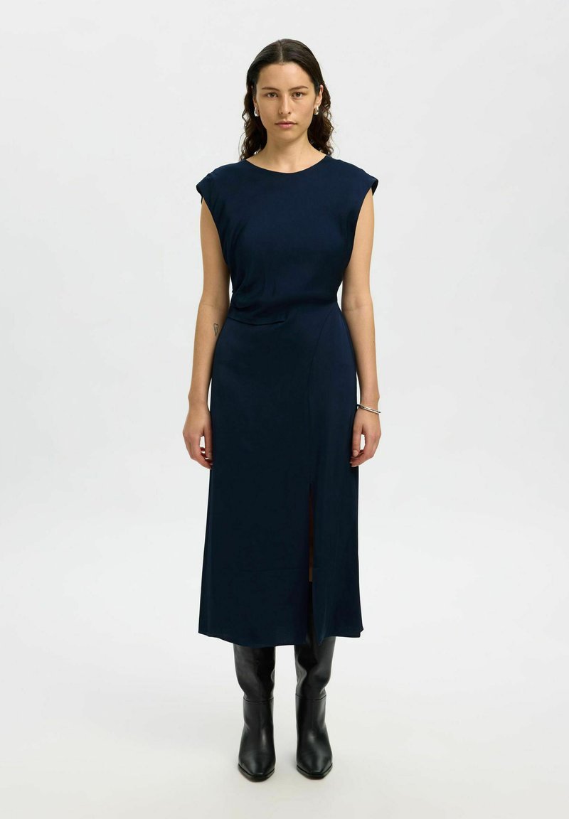 Femme portant une robe midi sans manches bleu marine avec une fente frontale, des bottes noires à hauteur de genou et des boucles d'oreilles en argent, se tenant devant un fond blanc uni.