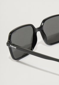 Μαύρα ορθογώνια γυαλιά ηλίου με σκούρους φακούς. Διαθέτουν γυαλιστερό πλαστικό σκελετό και λεπτό βραχίονα που εμφανίζει το λογότυπο "Polaroid".