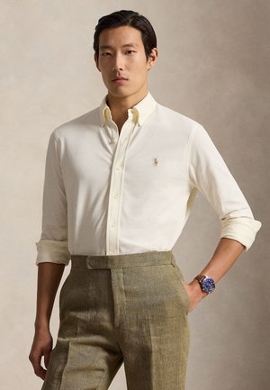 Homme portant une chemise crème à boutons avec logo, un pantalon marron taille haute et une montre bracelet au cadran bleu avec un bracelet en cuir.
