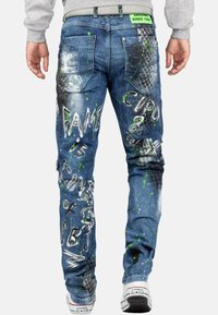 Jean denim bleu avec des détails usés, ornés d'accents peints en blanc et vert, et de texte. Équipé de cinq poches.