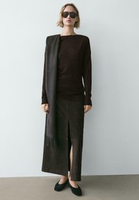 Brązowa bluzka z długim rękawem, ciemnobrązowa spódnica z bocznym rozcięciem oraz ciemny szal przewieszony przez jedno ramię, do tego czarne płaskie buty.