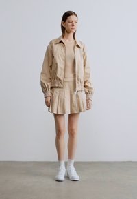 Beige bomberjack met elastische manchetten, geribde beige top en geplooide beige rok. Gecombineerd met witte sneakers en enkellange witte sokken.