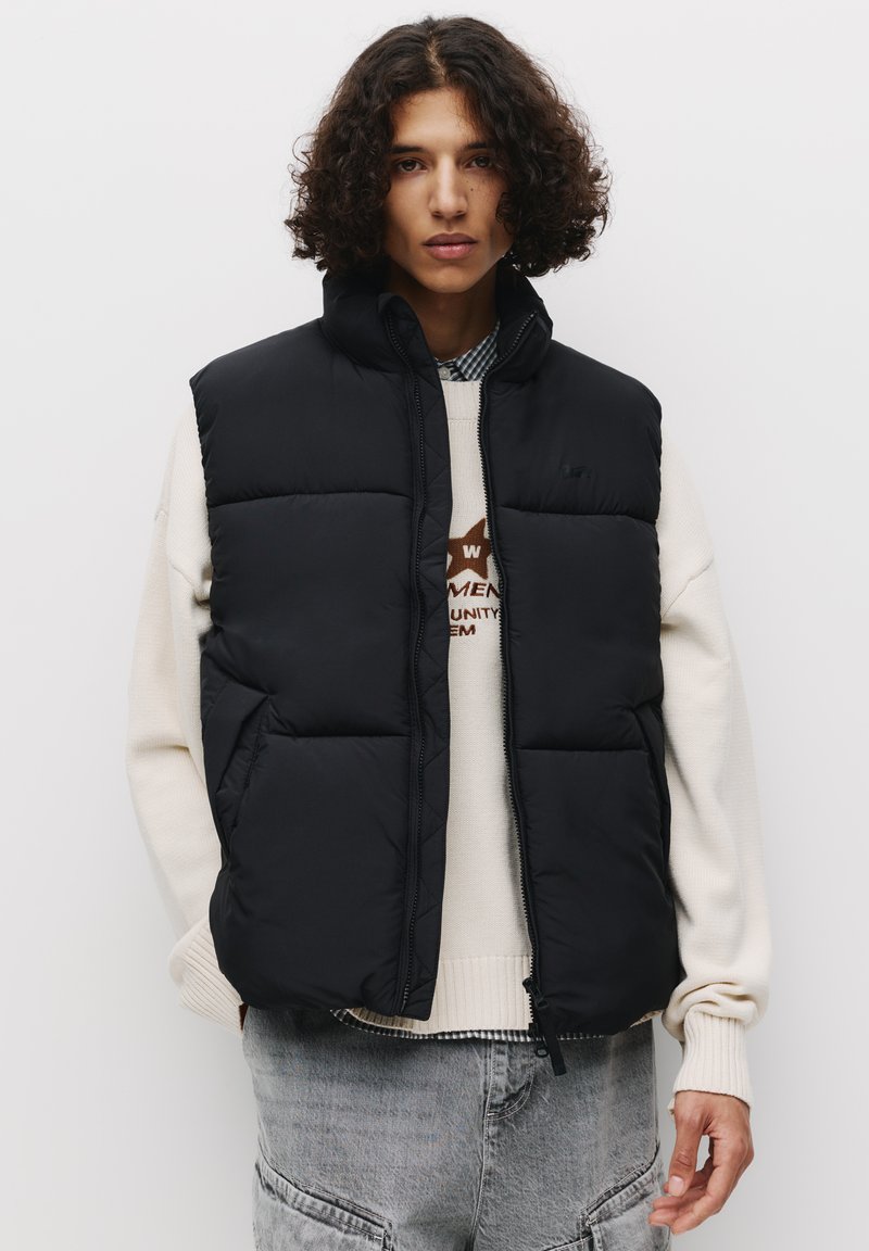 Gilet piumino nero con colletto alto, chiusura con zip e design trapuntato. Indossato sopra un leggero felpa con un motivo grafico.