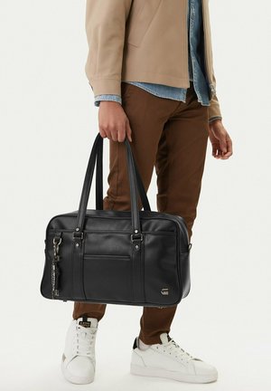 Person in brauner Hose und weißen Turnschuhen hält eine schwarze Lederhandtasche mit doppelten Griffen und Fronttasche.