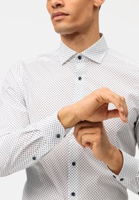 Chemise blanche à pois noirs, col pointu, boutons bleus, manches longues avec poignets à boutons, texture de tissu lisse.