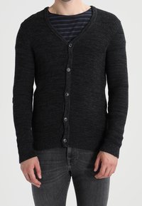 Svart stickad kofta med V-hals, med sex knappar framtill och ribbade muddar. Bärs över en marinblå randig skjorta.