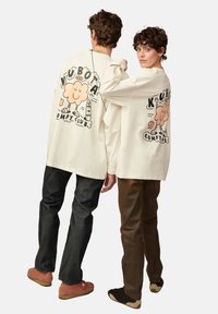 Two individuals wear cream oversized long-sleeve shirts with cartoon graphics on the back. One pair of dark jeans, one tan pants, mixed shoe styles.