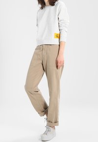 Jasnoszara bluza, okrągły dekolt, z żółtą naszywką logo CK. Beżowe spodnie tapered z podwiniętymi mankietami. Białe sneakersy dopełniają stylizację.