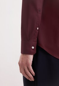 Camicia bordeaux a maniche lunghe con polsini con bottoni, caratterizzata da un tessuto testurizzato e un piccolo dettaglio ricamato a forma di rosa sul polsino.