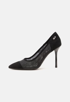 Sort pumps med høj hæl med mesh sidepaneler, ruskindståhæt og "Karl" broderi nær hælen på hvid baggrund.