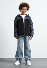 Padded jakna s gornjim dijelom u mornarskoj plavoj boji, crni fluffy fleece u sredini i patentnim zatvaračem. Nosena preko bijele košulje, u kombinaciji s svijetloplavim trapericama.