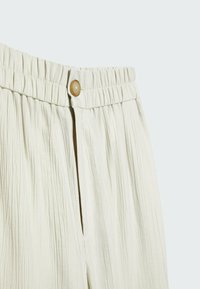 Gros plan sur un pantalon de couleur crème avec une ceinture élastique, des plis verticaux et une fermeture à un seul bouton marron clair à l'avant.