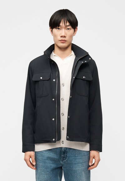 Jeune homme portant une veste noire zippée avec des poches poitrine boutonnées, par-dessus un cardigan blanc et un jean bleu, debout devant un fond uni.