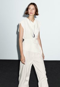 Blouse blanche à manches courtes avec un col drapé, associée à un pantalon léger beige ample. Un long collier en argent avec un pendentif ajoute une touche de détail.