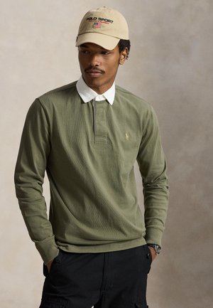 Homme portant une chemise à manches longues vert olive avec col blanc, casquette Polo Sport beige et pantalon noir, debout les mains dans les poches.