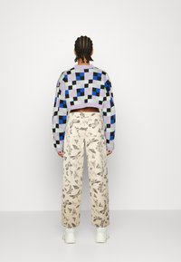 Sweater de crochê em padrão geométrico roxo, azul e verde; calças creme com estampas de borboletas cinzentas; modelo a usar ténis brancos.