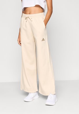 Personne portant un pantalon de survêtement beige à jambes larges avec taille à cordon et des baskets blanches, debout avec une main dans la poche.