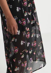 Käsi lepää lähellä läpinäkyvää mustaa mekkoa, jossa on värikkäitä pieniä kukkakuoseja violetissa, vaaleanpunaisessa, punaisessa, valkoisessa ja vihreässä.