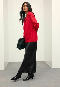 Cardigan rouge avec des boutons à l'avant, associé à une jupe noire en satin. Bottines noires à talons pointus et un sac à main noir texturé.